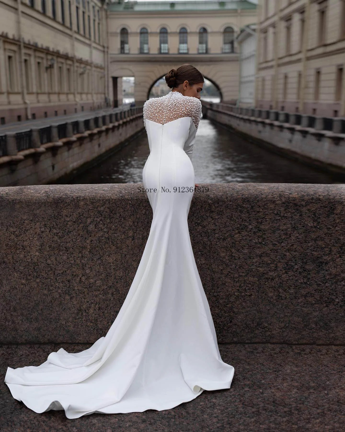 Bride in elegant white gown with beaded top stands on a stone bridge overlooking a narrow canal flanked by historic buildings. Her hair is styled in an elegant bun.