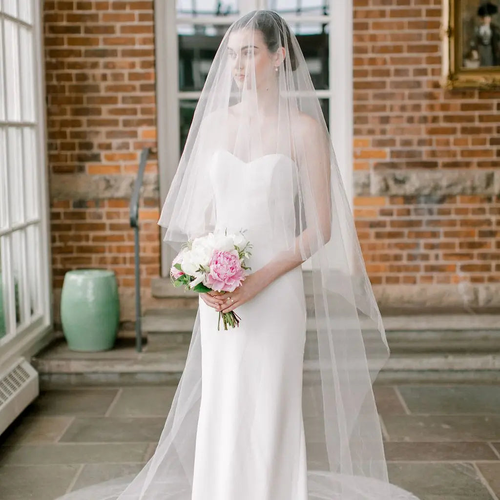 A bride in a strapless white gown and veil holds a bouquet of white and pink flowers, standing in a sunlit room with brick walls and large windows.