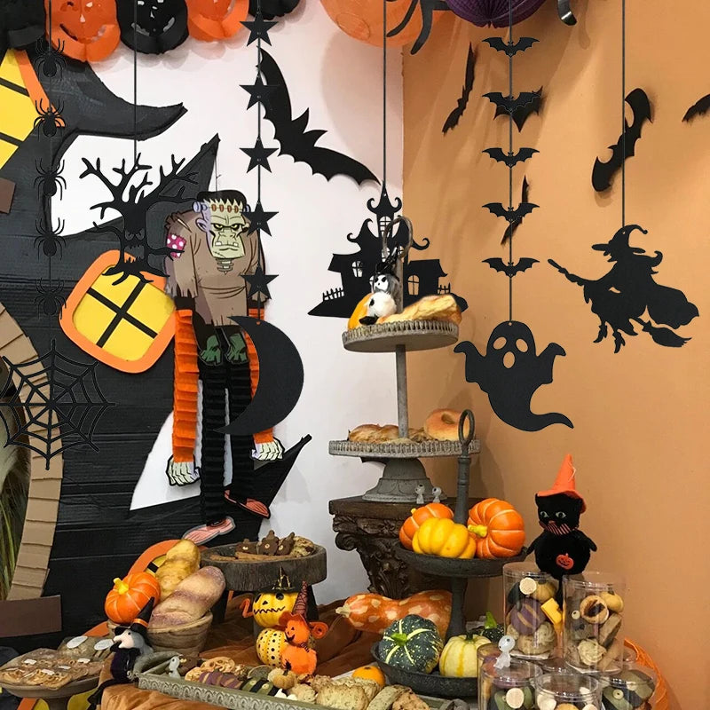 Halloween-themed display with black silhouettes of bats, a witch, and ghosts against a decorated wall. A tiered stand holds assorted pastries. Small pumpkins and festive figurines, including a black cat and a witch, are scattered on the table. Bright orange accents enhance the spooky atmosphere.