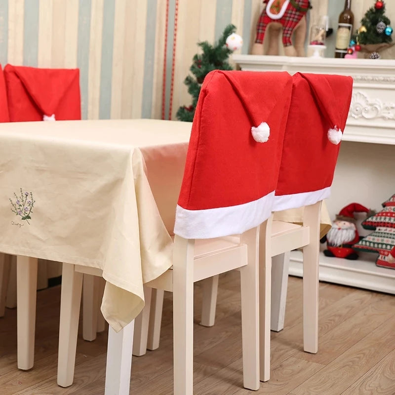 Dining chairs with Santa hat covers around a table with a cream tablecloth. Festive decor, including a small Christmas tree and Santa figurines, adorns the room.