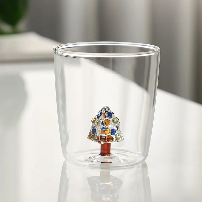 Double-walled glass cup with a colorful Christmas tree design inside, featuring blue, green, and orange dots. The cup is placed on a reflective white surface.