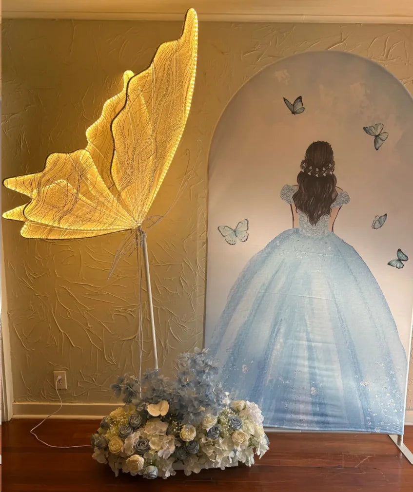 Illuminated butterfly wings stand next to a mural of a girl in a blue dress with butterflies around her. The base of the wings is adorned with white and blue flowers.