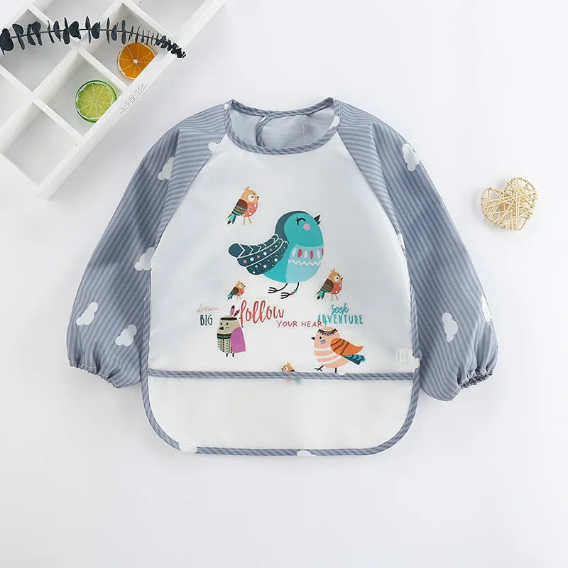 Children’s bib with long gray striped sleeves featuring white clouds, and a colorful bird design with playful text. Nearby are decorative items including citrus slices and a woven heart.