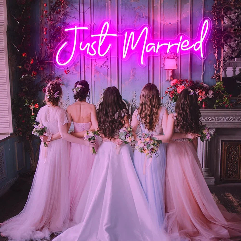 Four women in pastel gowns stand with backs turned, holding bouquets, facing a wall with a pink neon sign reading "Just Married." The setting is adorned with flowers.