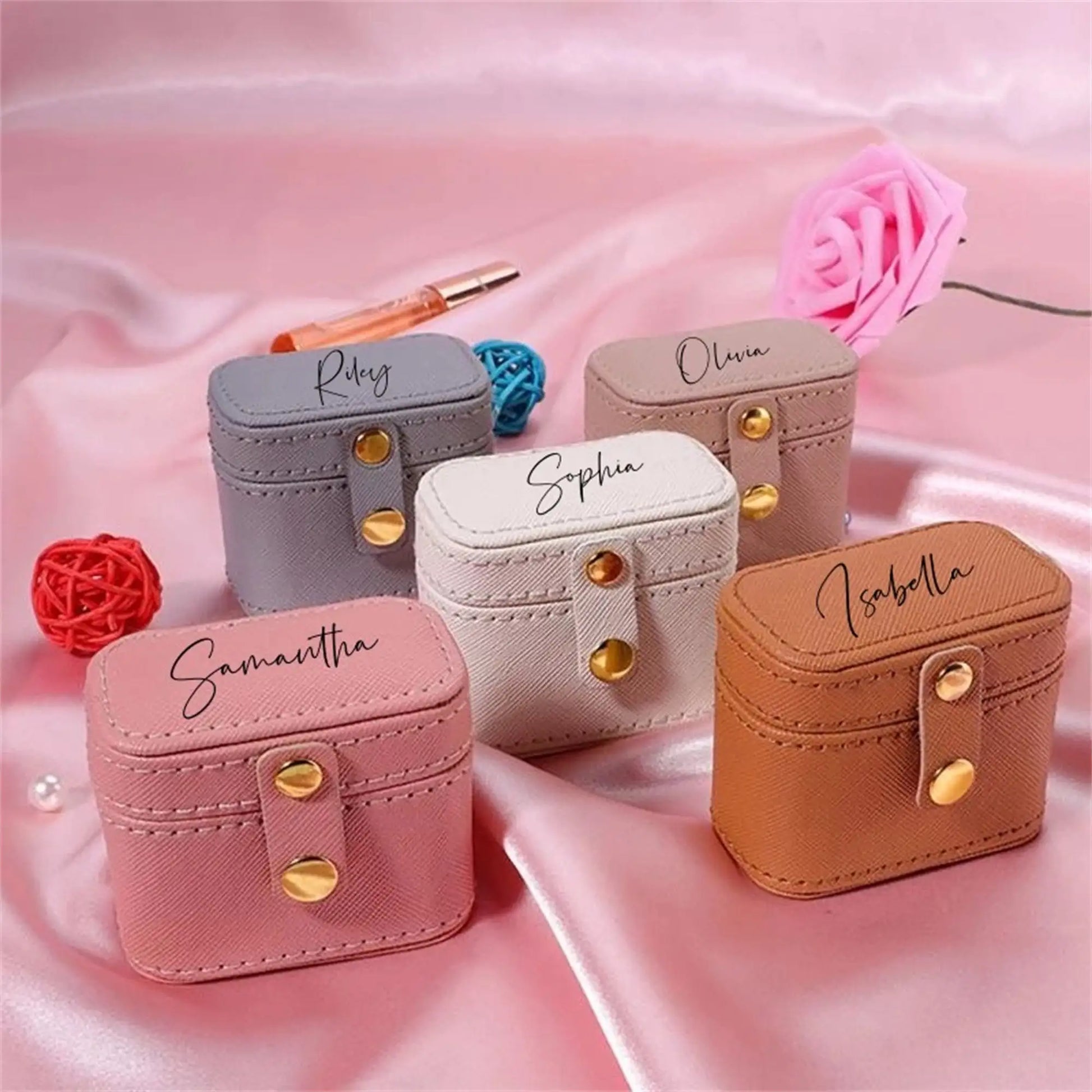 Five small, colorful leather pouches with names on top sit on pink satin fabric. Each pouch has a snap closure, surrounded by decorative balls and a pink rose.