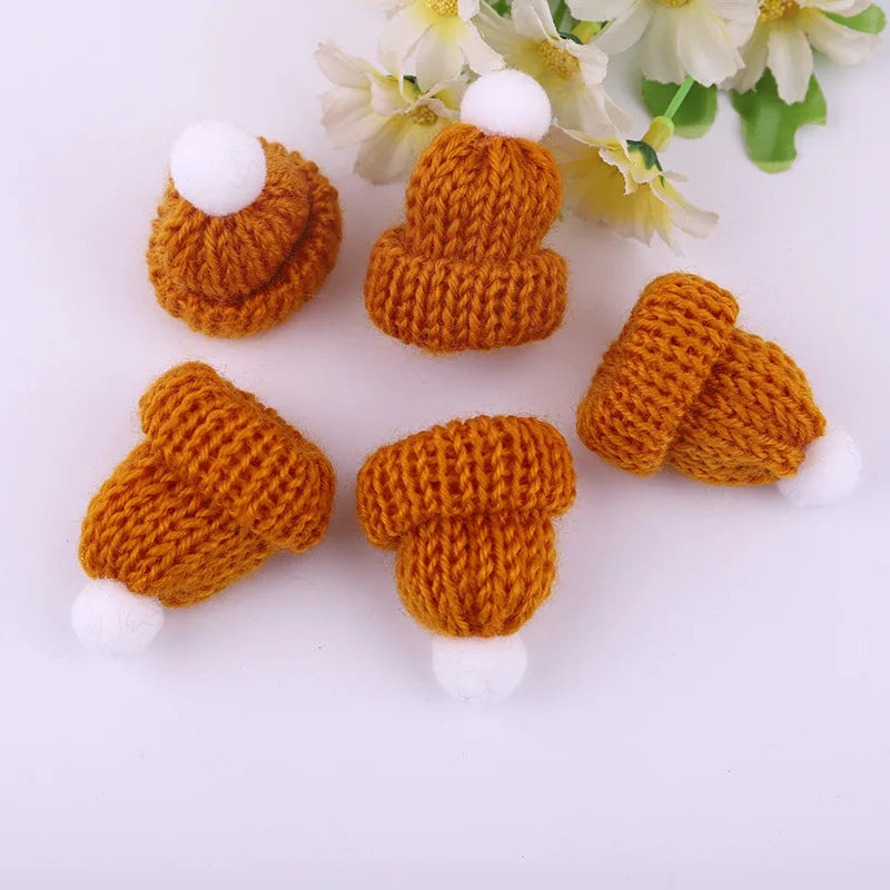 Five small, knitted orange beanies with white pom-poms are arranged on a white surface next to cream-colored flowers.