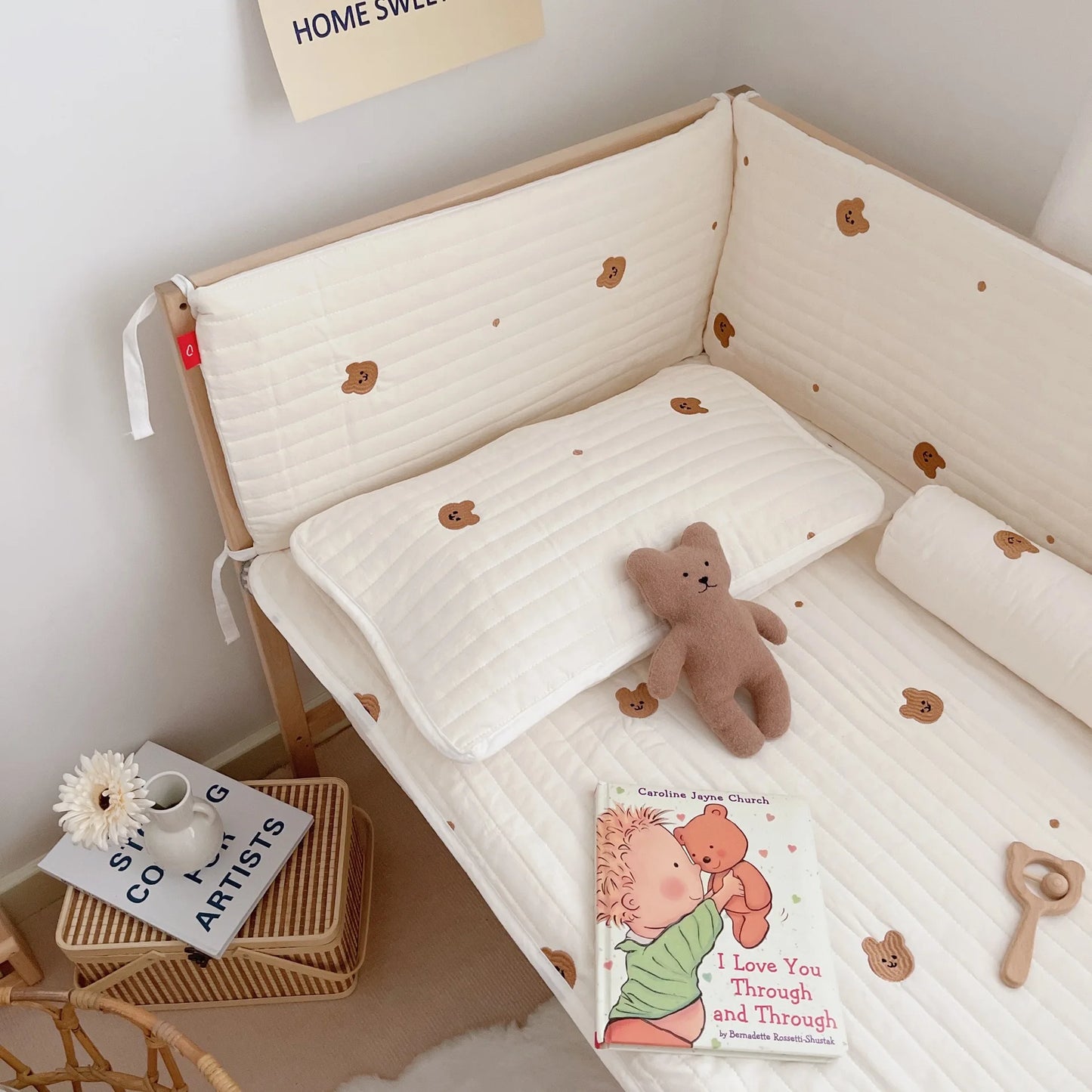Cozy baby crib with teddy bear bedding, a plush toy, and a wooden rattle. Nearby is a small table with a book titled "I Love You Through and Through" and a white vase with flowers.