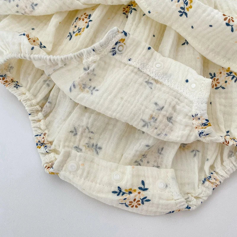 Close-up of a floral-patterned baby romper in soft fabric. It features a cream background with blue and yellow flowers, elastic leg openings, and snap closures at the bottom.