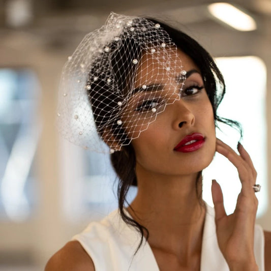 Woman with red lipstick wearing a white dress and a netted fascinator adorned with pearls. She stands in a softly blurred indoor setting, gently touching her face.