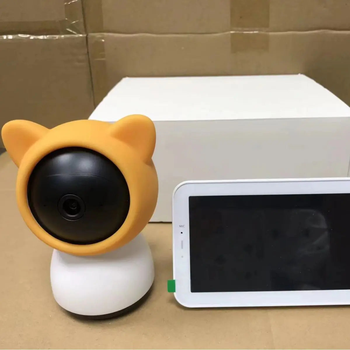 A security camera with bear ears beside a small rectangular black screen device on a cardboard surface with a white box in the background.