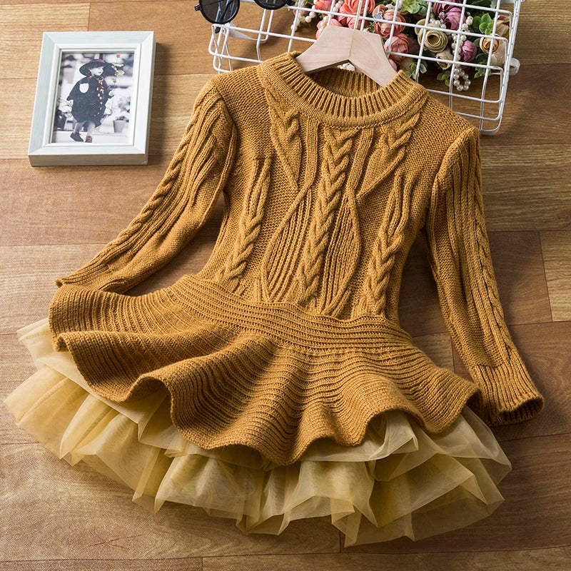 A mustard knit dress with a flared, layered tulle skirt lies on a wooden floor. Nearby, a framed black-and-white photo, sunglasses, and a basket with flowers add detail.