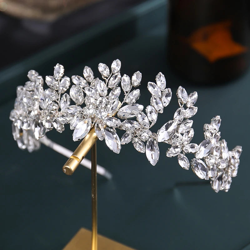 A sparkling silver tiara featuring clusters of marquise and round-cut crystals arranged in a floral pattern, displayed on a gold stand against a dark background.