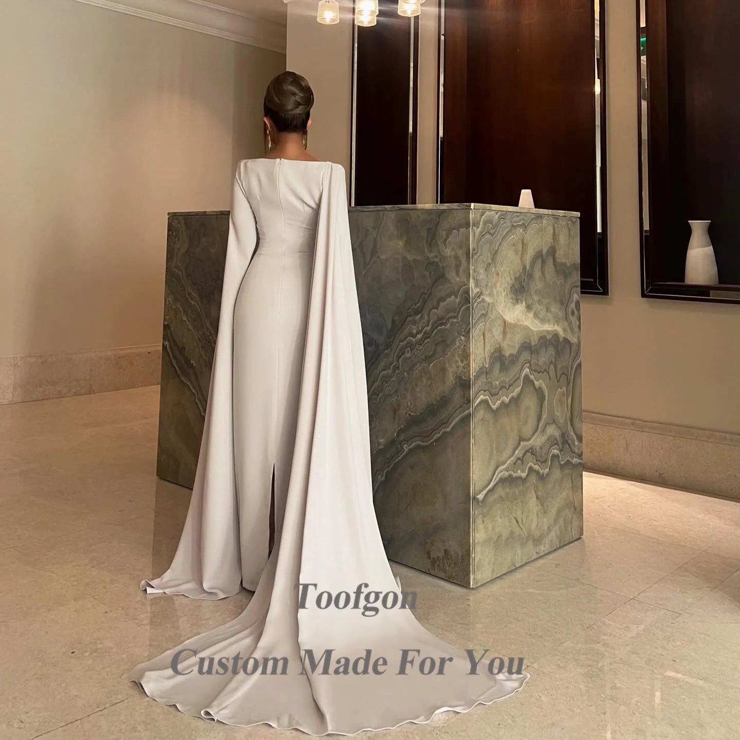 Elegant woman in a long, flowing white dress with a train stands at a marble reception counter, her back to the camera. The room has modern decor and soft lighting.