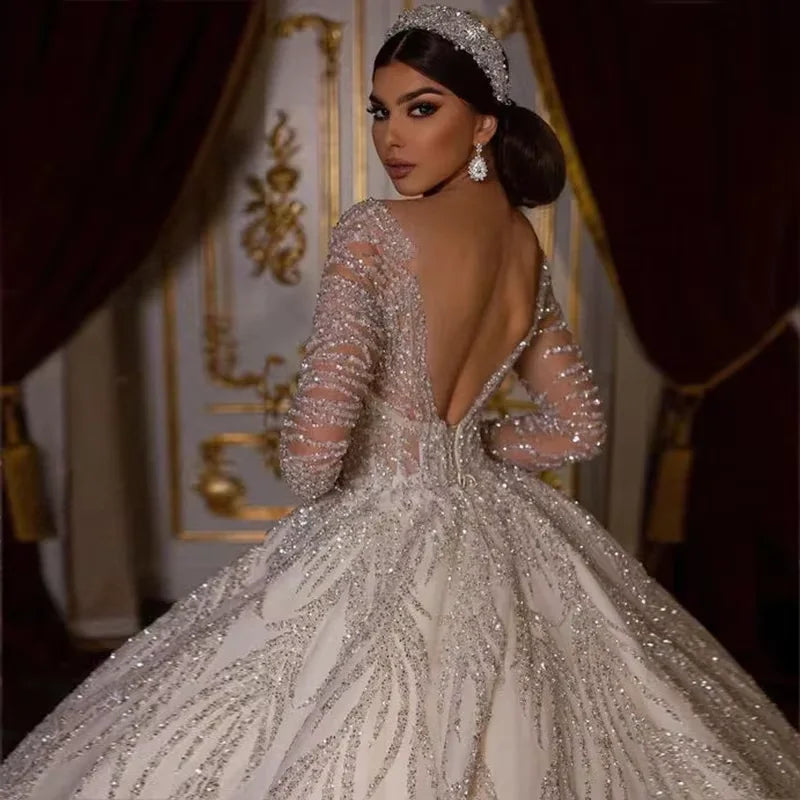 Bride in an elegant ball gown with intricate silver embellishments, posing with an elegant updo and sparkling earrings, stands against an ornate backdrop with rich drapery.