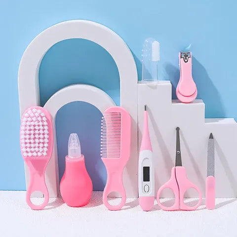 A pink baby grooming kit displayed against a blue background, including a hairbrush, nose aspirator, comb, thermometer, toothbrush, nail clippers, scissors, and file.