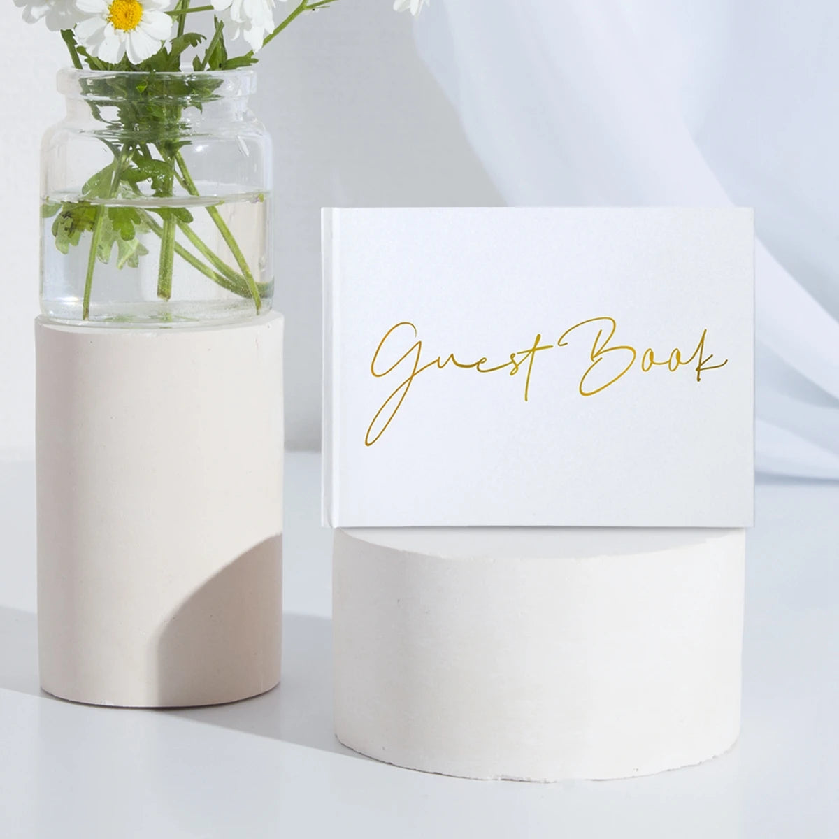 White guest book with gold script placed on a round pedestal. A glass jar with white daisies sits beside it, and both are set against a soft, light background.