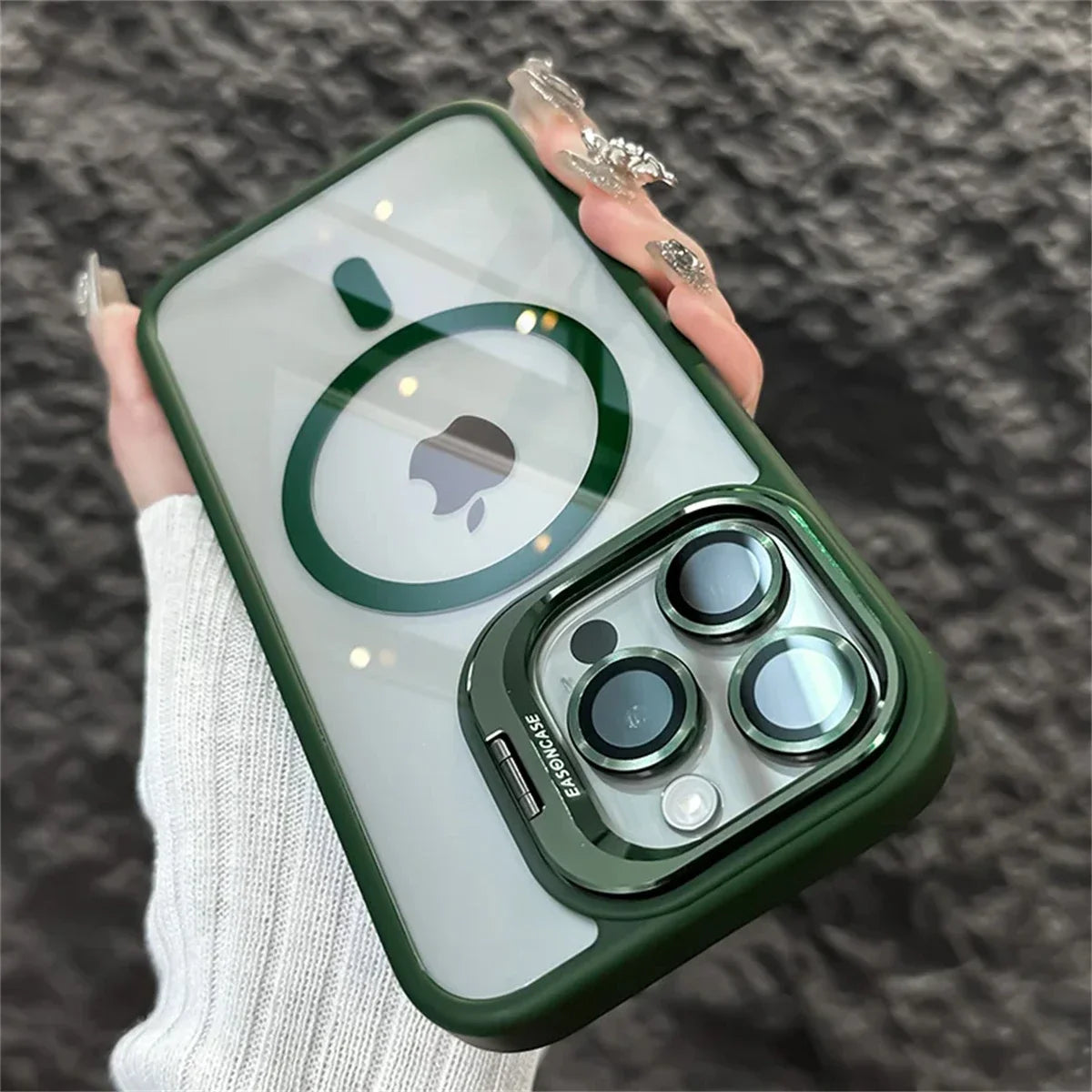 A hand holds a smartphone with a green and clear case, featuring a visible Apple logo and a prominent camera design. The person's nails are decorated with intricate designs.