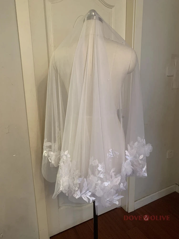 A sheer white veil featuring delicate floral embroidery along the edges is draped over a dress form, set against a plain door background. The floor is wooden.
