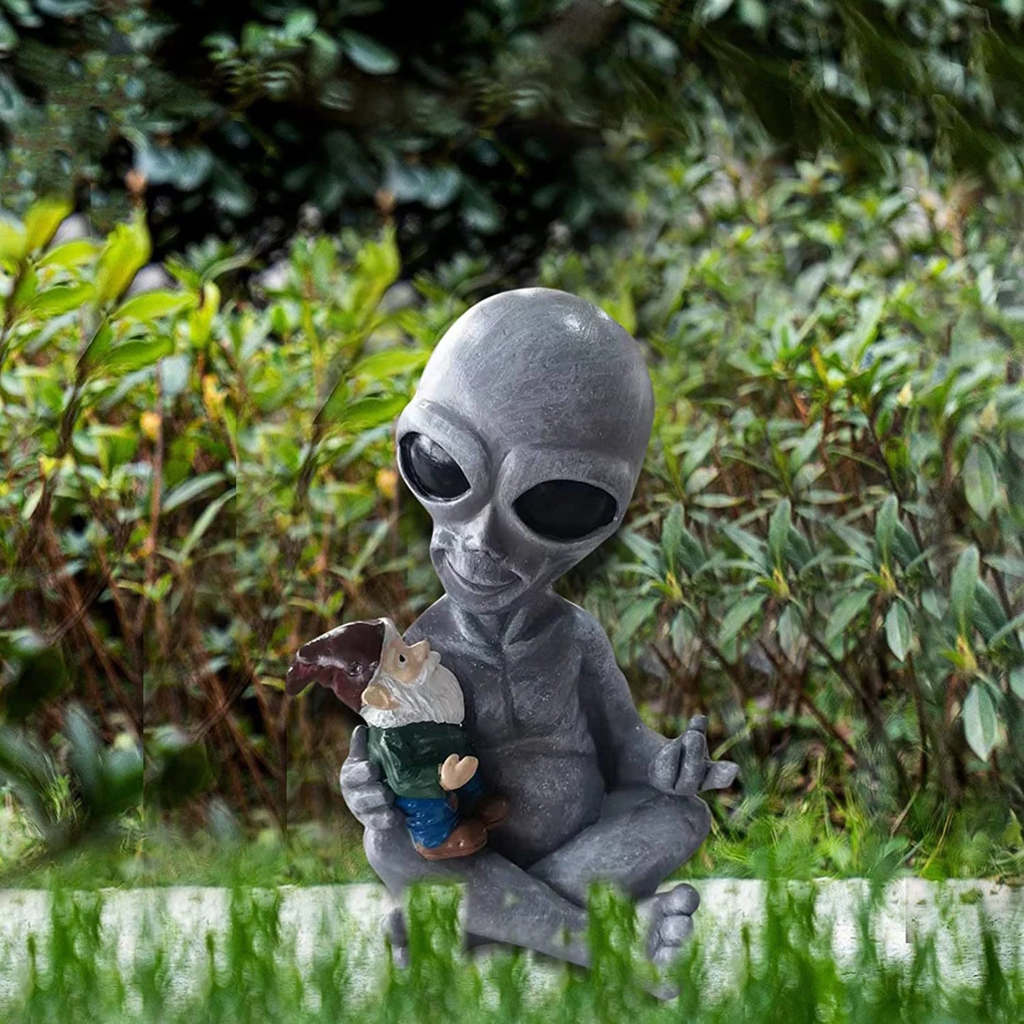 A grey alien statue with large eyes holding a small garden gnome sits cross-legged in a lush garden, surrounded by green foliage and grass.
