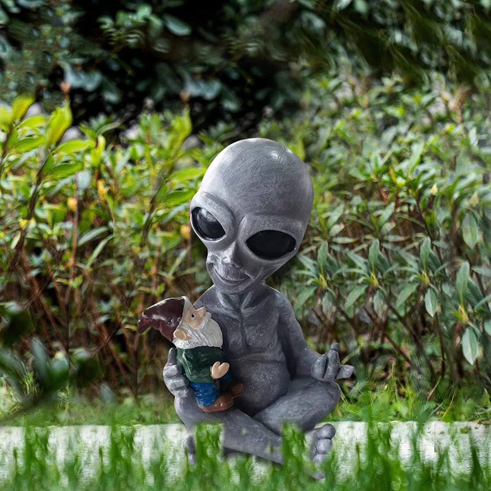 A garden statue of a seated alien with large black eyes cradles a small bearded gnome. The scene is set against lush green foliage, adding a whimsical touch.