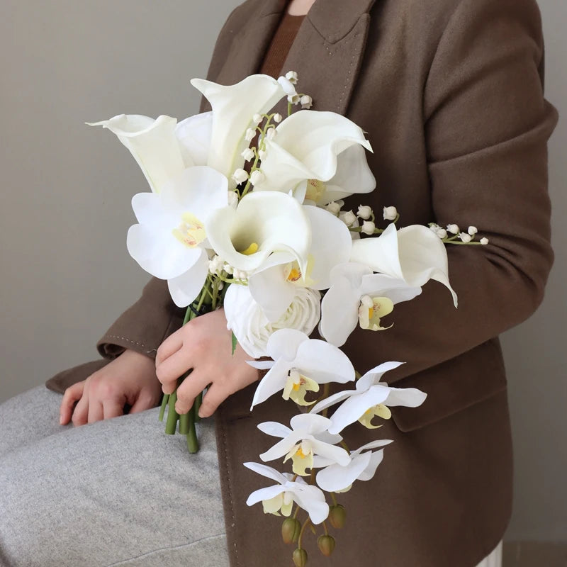 A person in a brown coat holds a bouquet featuring white calla lilies, orchids, and lily of the valley against a neutral background.