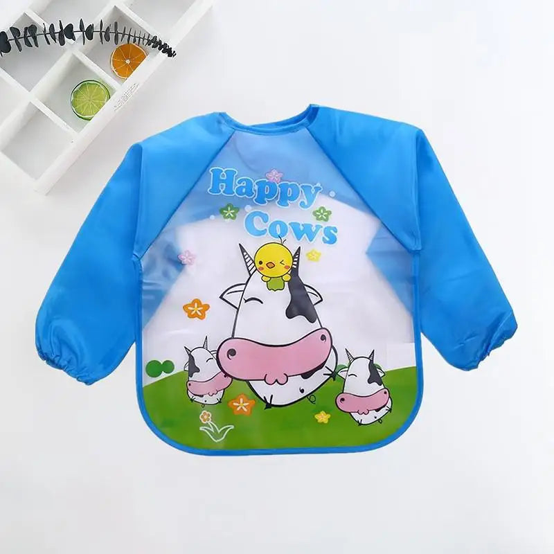 Blue long-sleeve bib featuring cartoon cows and a chick, with the text "Happy Cows." Background shows a grid shelf with decorative items.