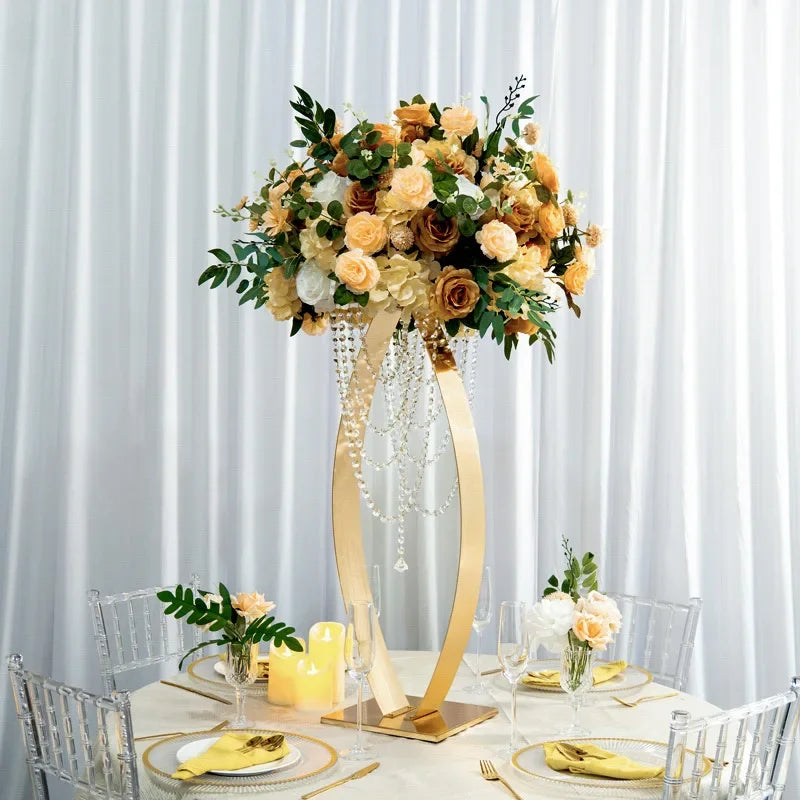 Elegant table setting with a tall floral centerpiece of peach and white roses, greenery, and hanging crystals. Surrounded by glassware, candles, and gold-accented dinnerware on a round table.