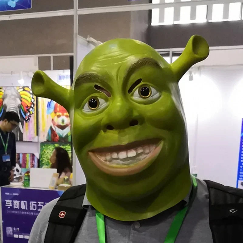 Person wearing a Shrek mask at an indoor event. Behind them, colorful artworks are displayed on walls. People are interacting at various booths in the background.