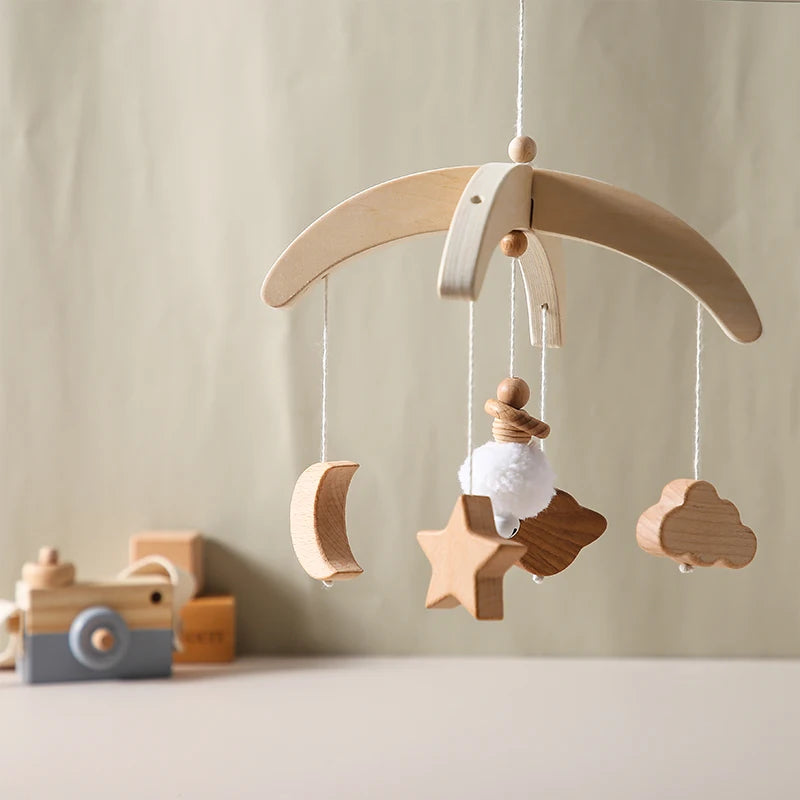 Wooden baby mobile with hanging crescent moon, star, cloud, and pompom sheep. Background features wooden blocks and a camera toy on a neutral surface.