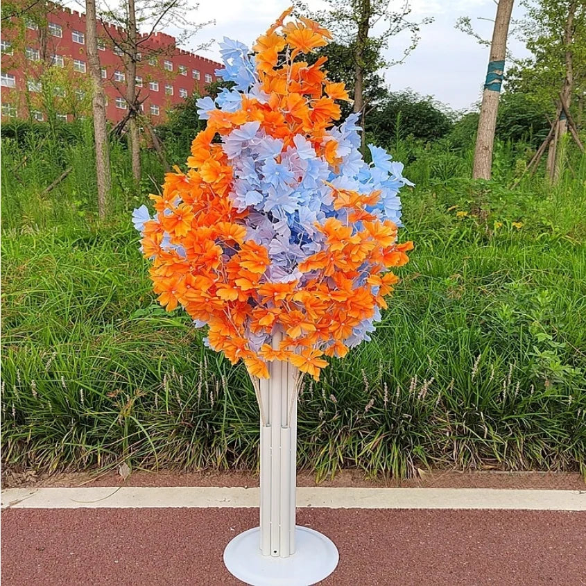 Artificial tree with vibrant orange and pastel blue flowers stands on a white base, set against a background of green grass and a red building.