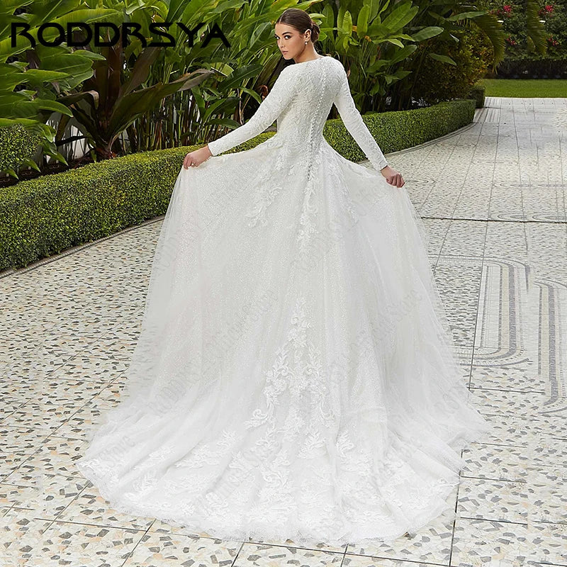Bride in a long-sleeve lace wedding gown holds her train, standing on a patterned stone path, surrounded by lush greenery.