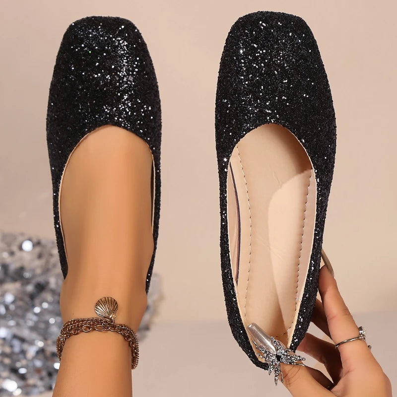 Black glittery ballerina flats worn on feet, one foot adorned with a shell anklet. Nails are manicured and shiny, against a neutral background with sparkling effects.