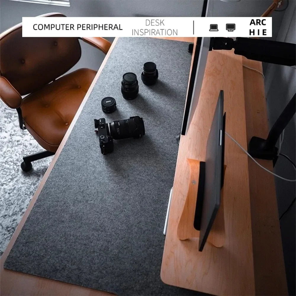 Minimalist desk setup with a gray mat, camera, and lenses. A sleek monitor is mounted on a wooden stand, beside a brown leather chair on a gray carpet.