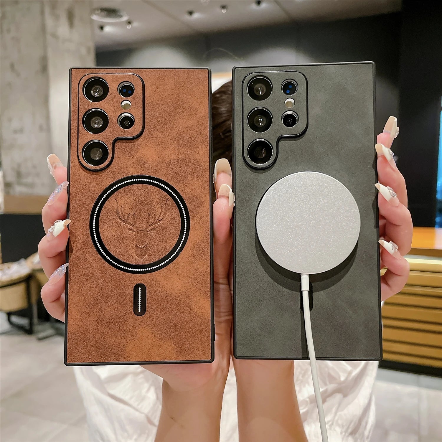Two phones are held side by side. The left phone has a brown leather case with a deer emblem, while the right phone has a black case with a round, silver charging pad attached.