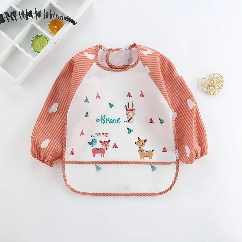 Children's bib with orange striped sleeves, cloud patterns, and a front design featuring animals, the words "Be Brave" and "Dream Big." Nearby, a decorative heart and shelf.