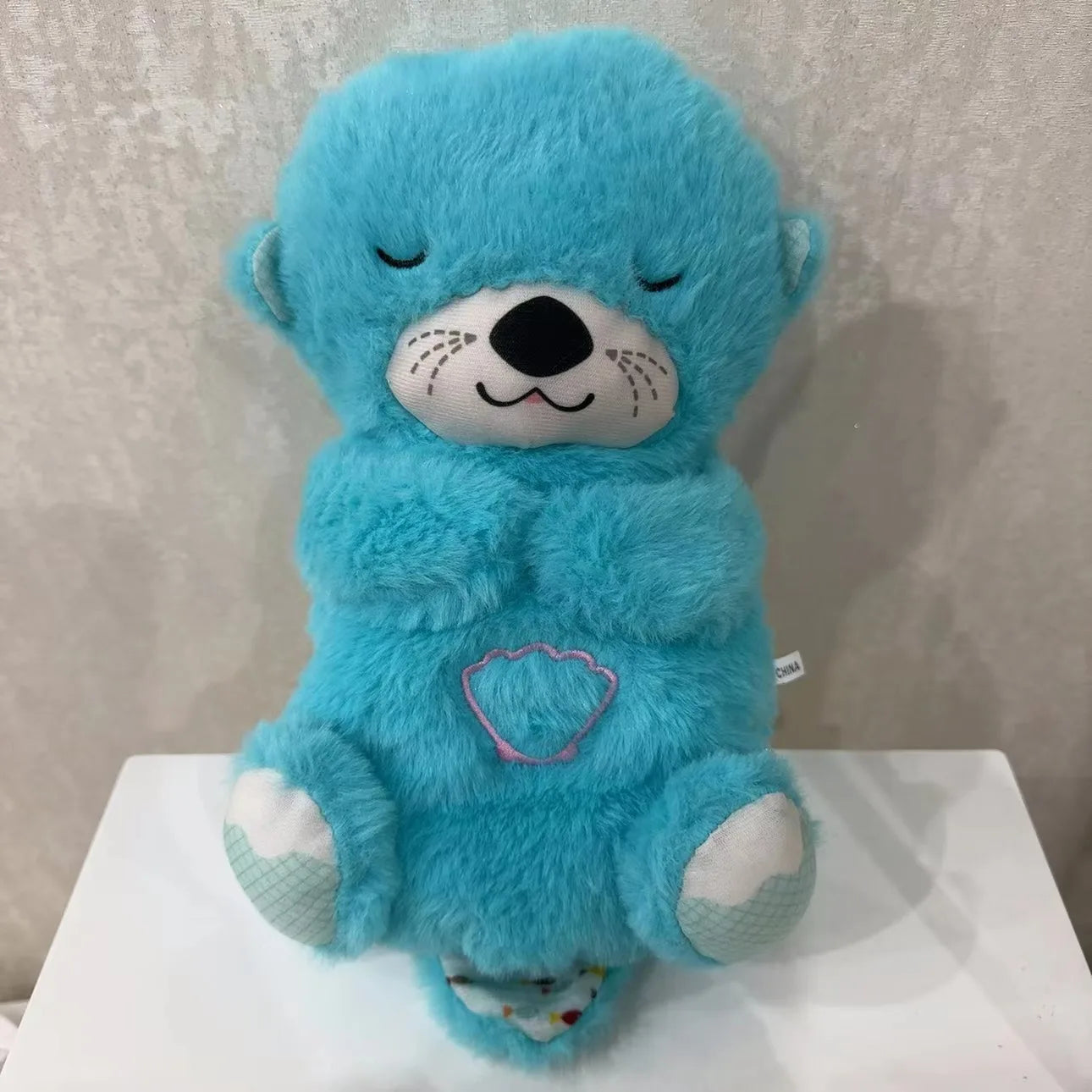 Plush blue otter with stitched eyes and nose, holding its paws together. It has a pink shell emblem on its belly and is set against a light background.