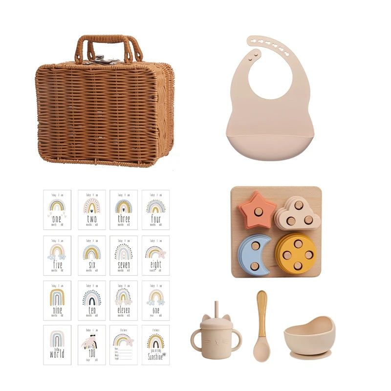 Wicker basket with baby essentials: beige bib, milestone cards with rainbows, wooden stacking toy, sippy cup with cat ears, spoon, and bowl.