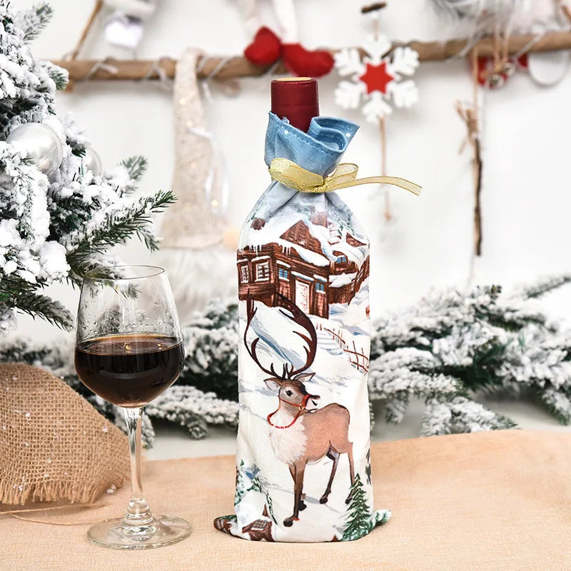 A wine bottle wrapped in holiday fabric with a reindeer and snowy cabin design sits in front of a glass of red wine, surrounded by festive, snow-covered greenery and decor.