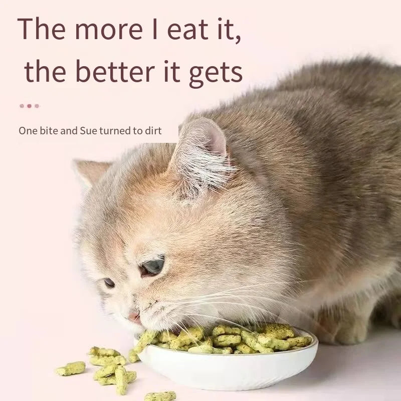A brown cat eagerly eats green cat treats from a white bowl on a pink background. Text above reads, "The more I eat it, the better it gets." Below, "One bite and Sue turned to dirt."
