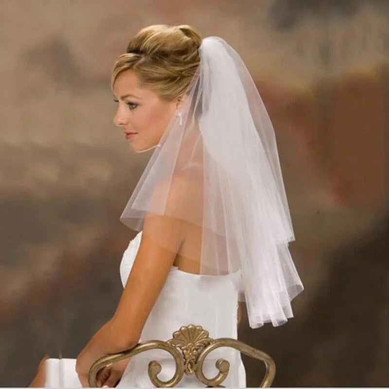 Bride with an elegant updo and delicate white veil sits on a decorative chair, wearing a strapless white dress, against a soft, blurred background.