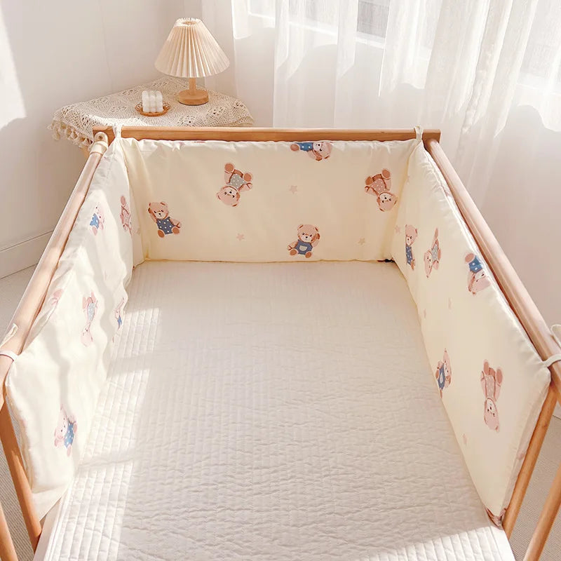 Crib with soft teddy bear-patterned bumpers, a quilted mattress, and a small bedside table with a decorative lamp by sheer curtained window.