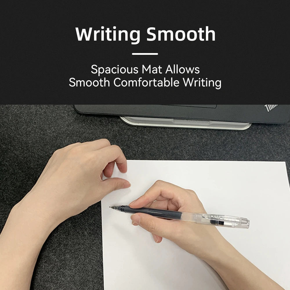 Hands writing on white paper on a dark mat. Text above reads: "Writing Smooth - Spacious Mat Allows Smooth Comfortable Writing." A laptop is partially visible in the background.