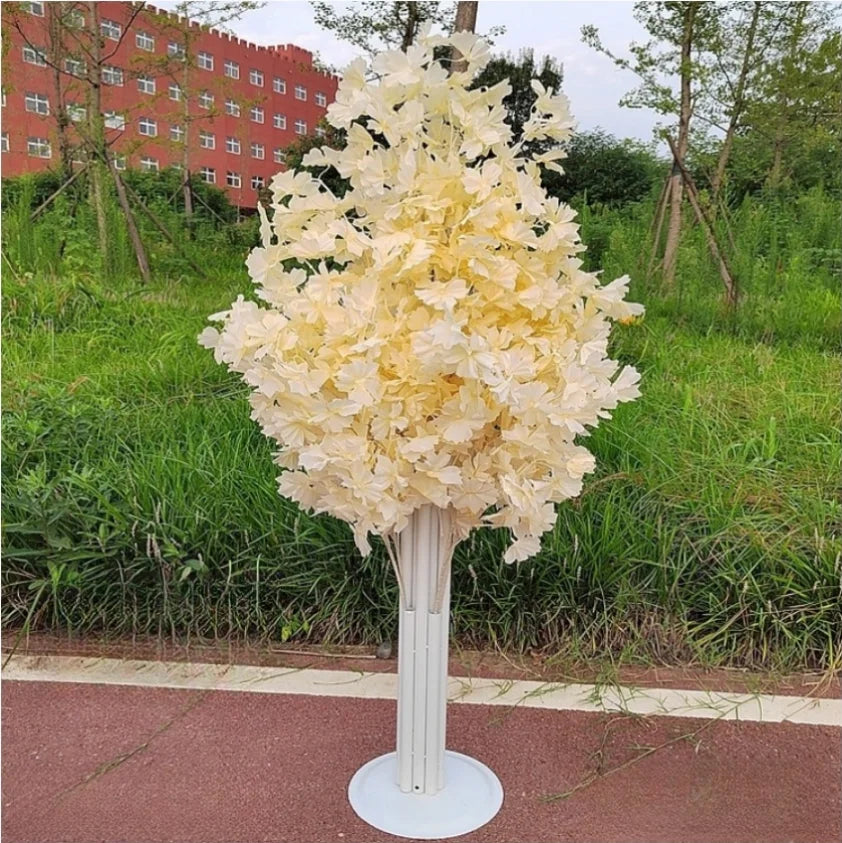 A decorative paper tree with cream-colored leaves on a tall, white stand set against a grassy background with a red building in the distance on a cloudy day.