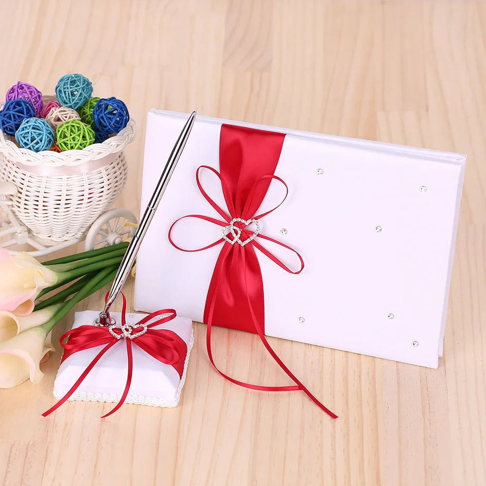White guestbook with red ribbon and heart embellishment, matching pen holder on wooden table. Basket of colorful wicker balls and flowers in background.