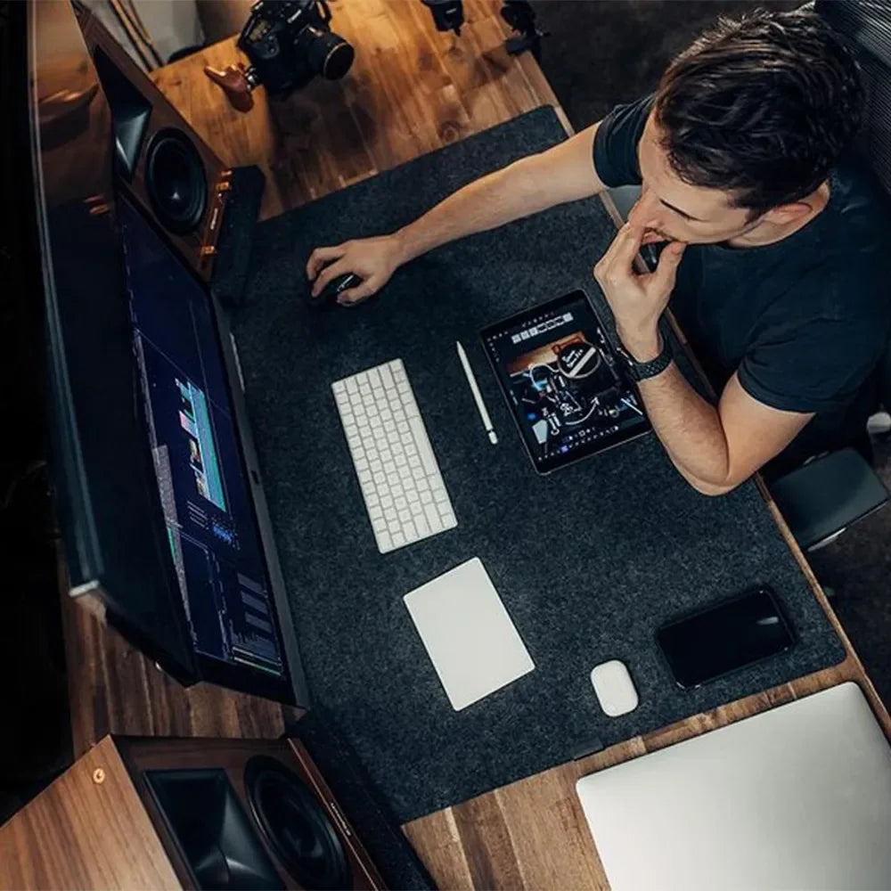 Man working at a wooden desk with a large monitor, keyboard, tablet, and smartphone. He is using a mouse, surrounded by tech gadgets and camera equipment.