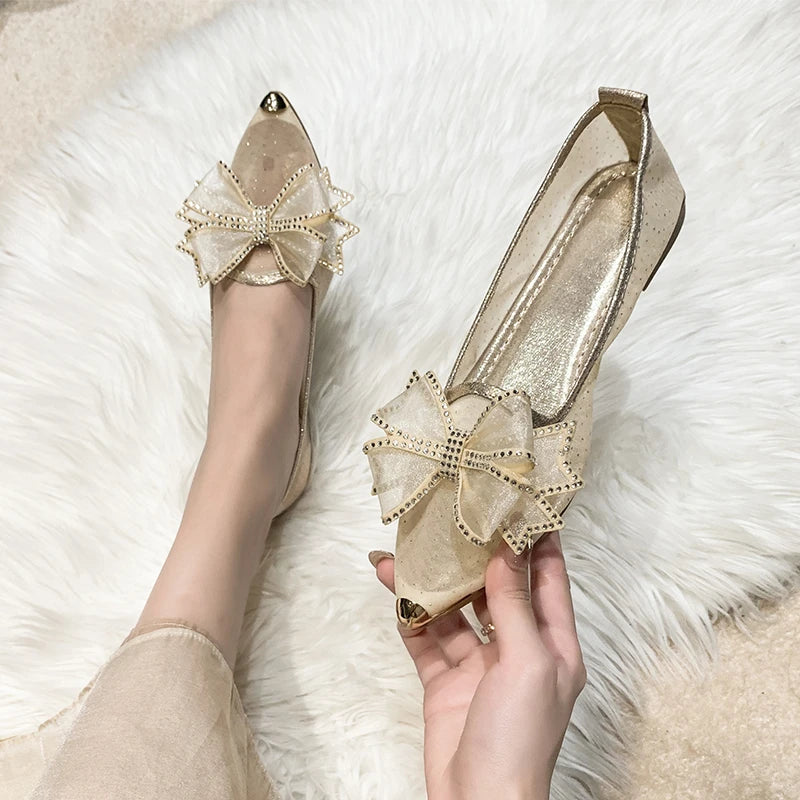 Elegant gold heels with transparent mesh, adorned with large bows and rhinestone accents, held in hand and worn on foot, placed on a fluffy white rug.