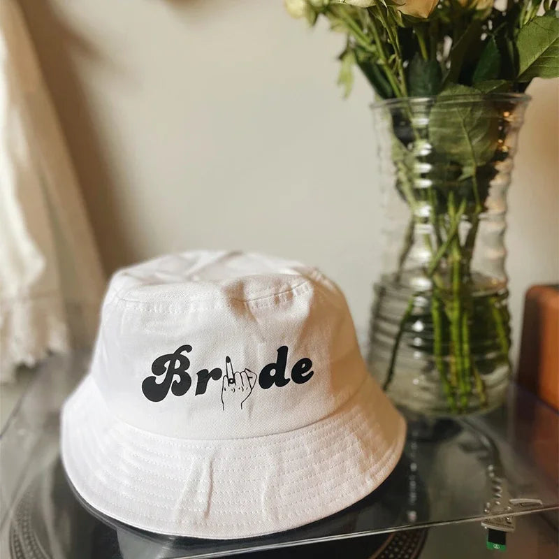 White bucket hat with the word "Bride" and a hand gesture, placed on a clear surface near a vase of white roses.