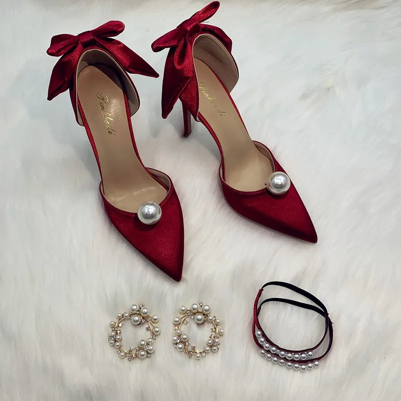 Red satin high heels with pearls and bows on a white surface, paired with pearl hoop earrings and a pearl bracelet, creating an elegant, coordinated look.