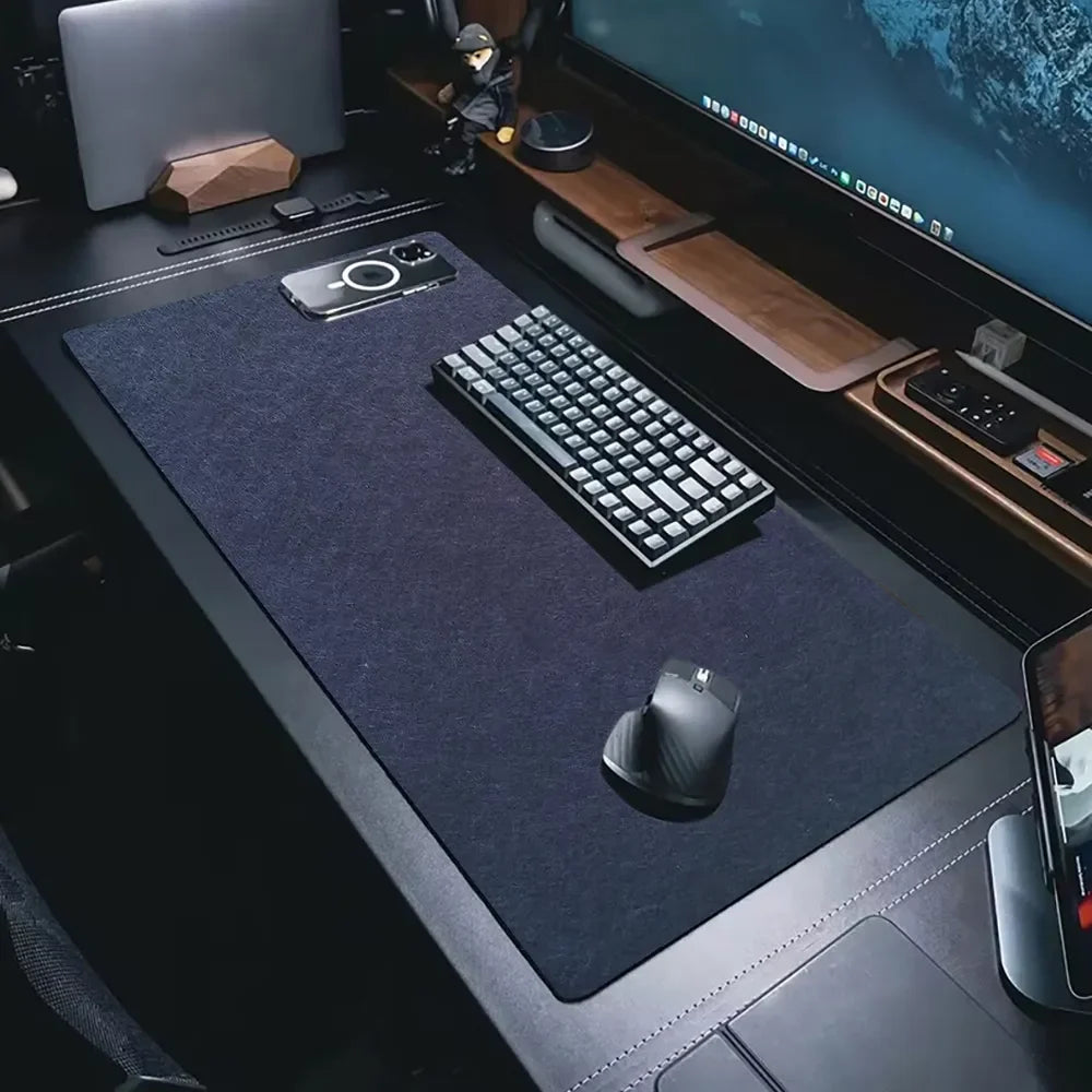 A tidy desk setup with a large black mat, mechanical keyboard, ergonomic mouse, and smartphone. A monitor, smartwatch, and figurine are on the wooden shelf in the background.