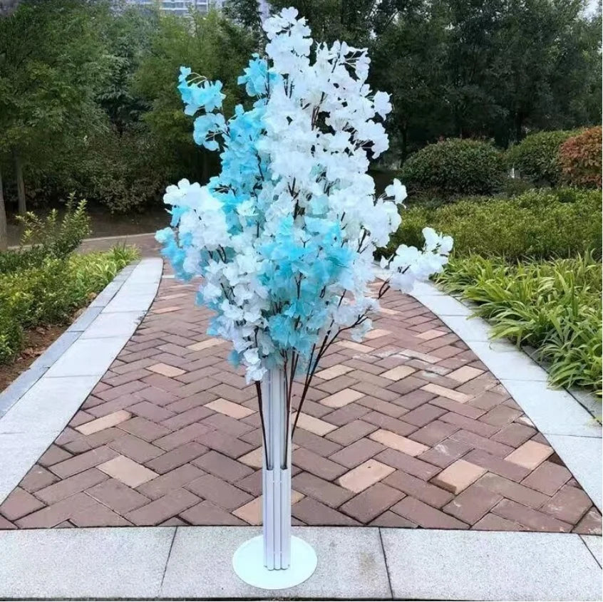 Artificial white and blue decorative flowers in a white vase are placed on a paved pathway, surrounded by lush green bushes and trees in a park setting.